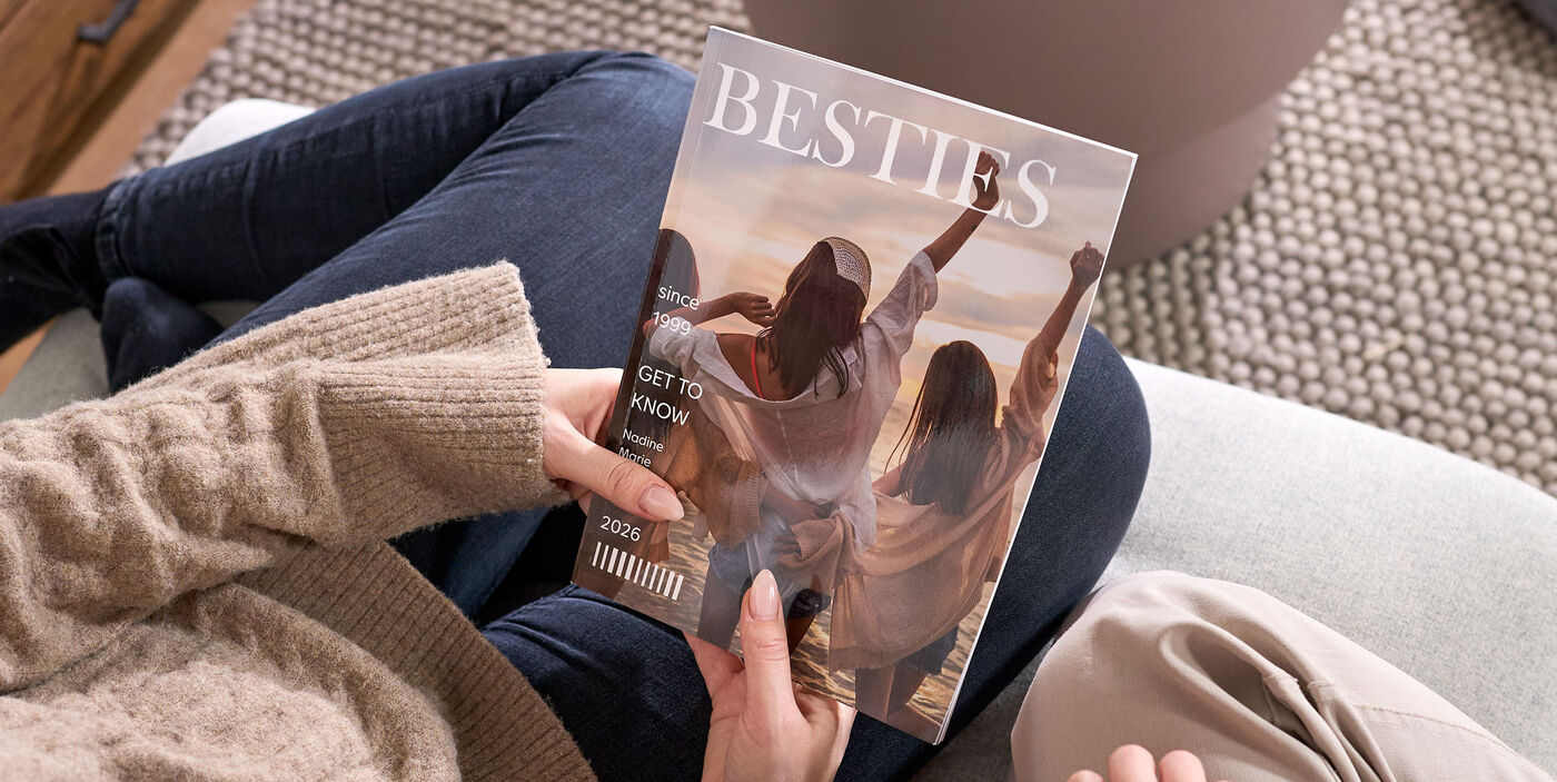 Două persoane stau pe canapea și privesc o revistă închisă, pe coperta căreia apar trei femei văzute din spate, pe plajă, alături de titlul „BESTIES”.